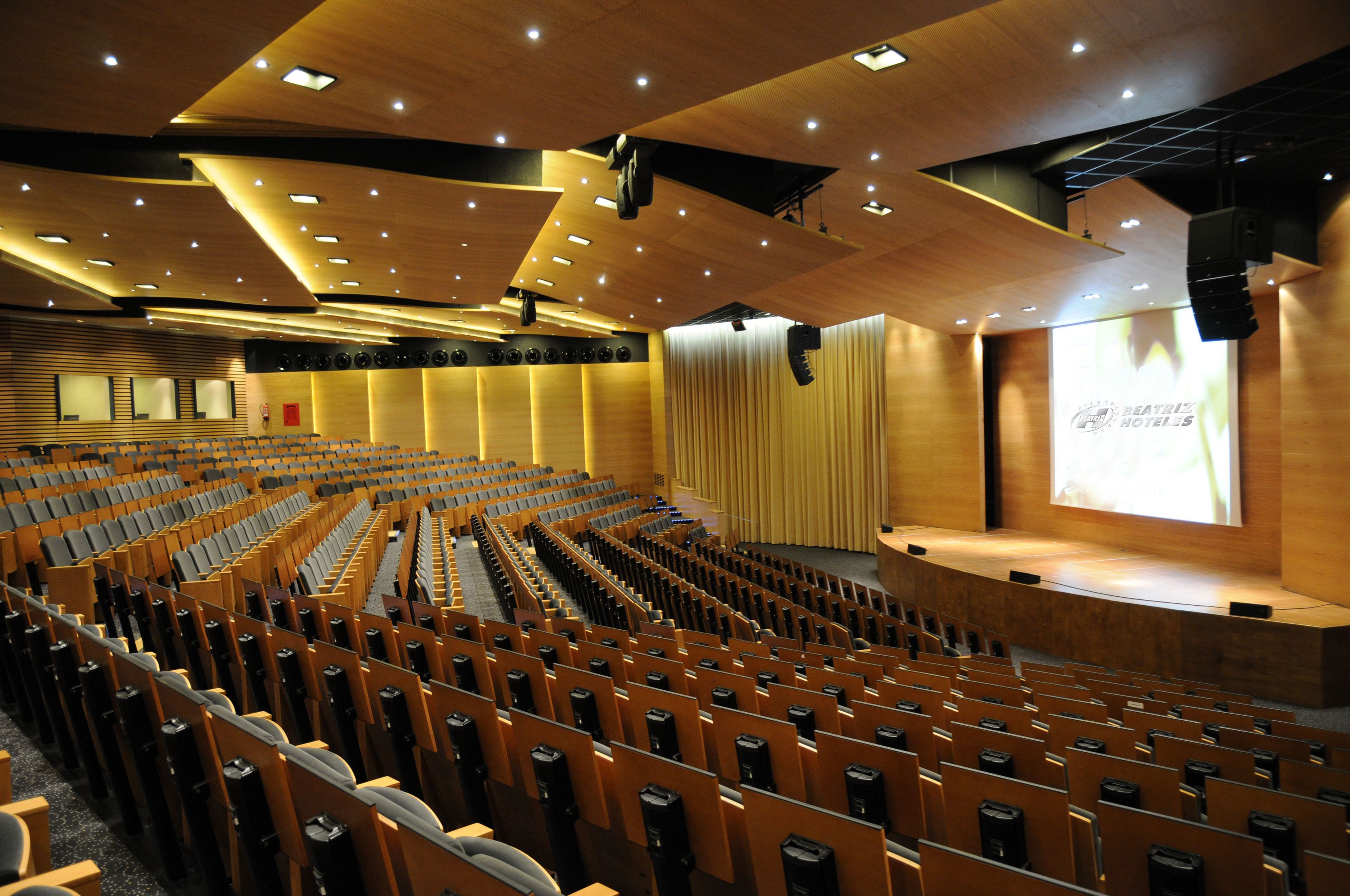 A large auditorium with rows of wooden seats, a stage, and a projection screen displaying 'Beatriz's Hotels'.