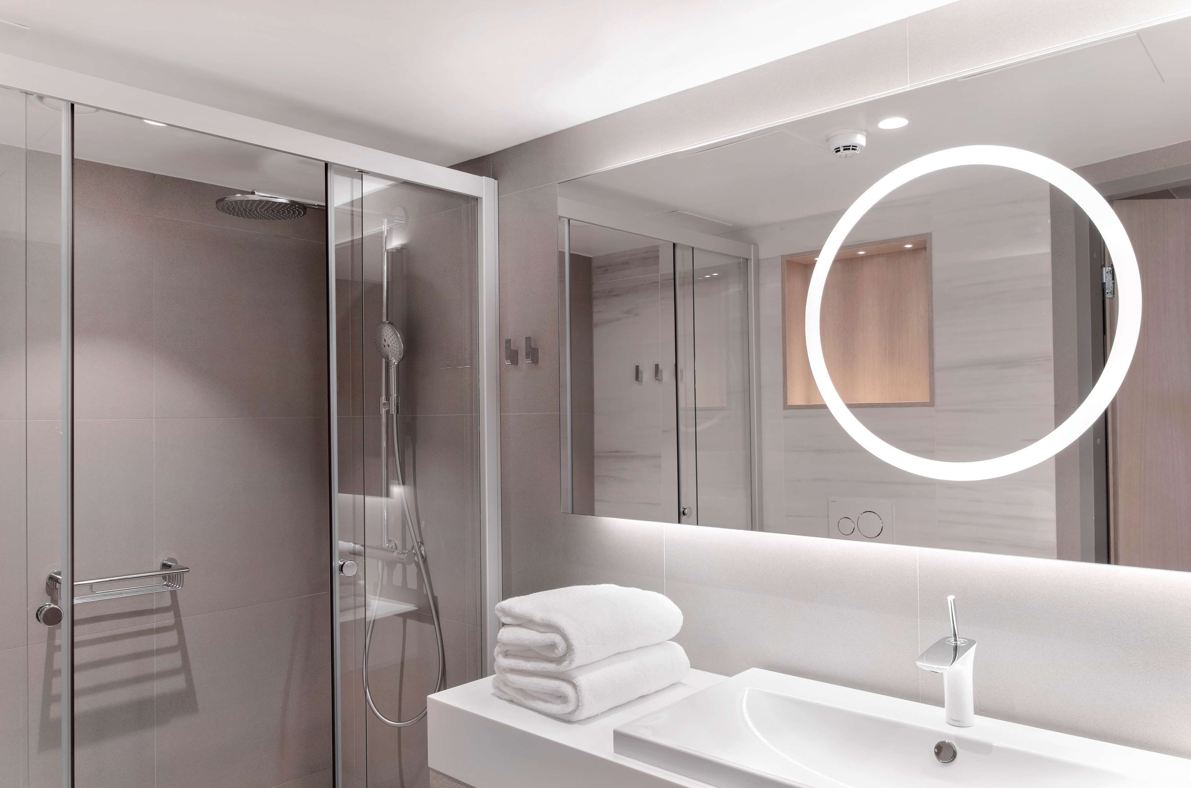 A modern bathroom with a glass-enclosed shower, a white sink, and a large mirror with a circular light.