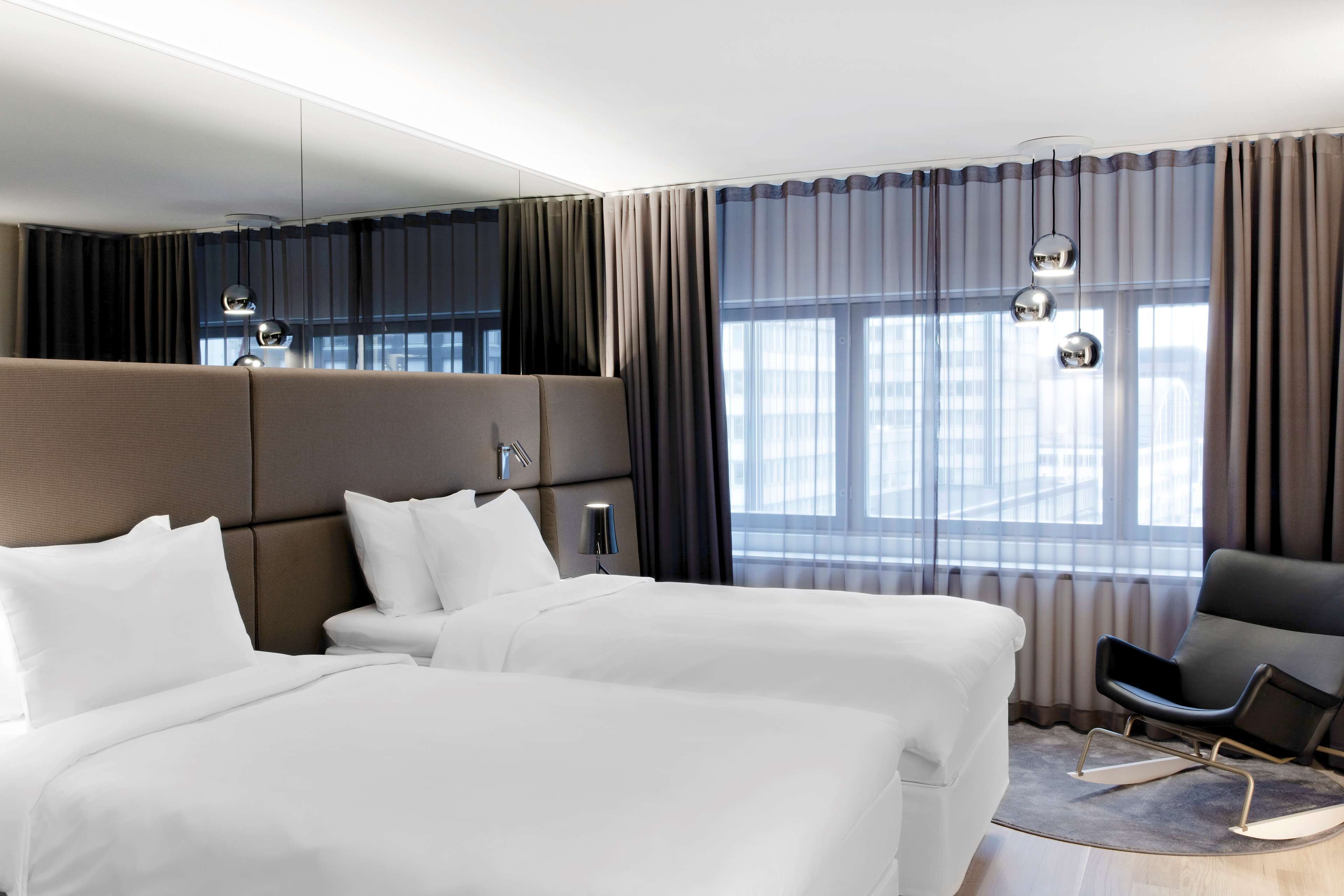 A hotel room with two beds, a chair, a round table, and a large window with curtains.