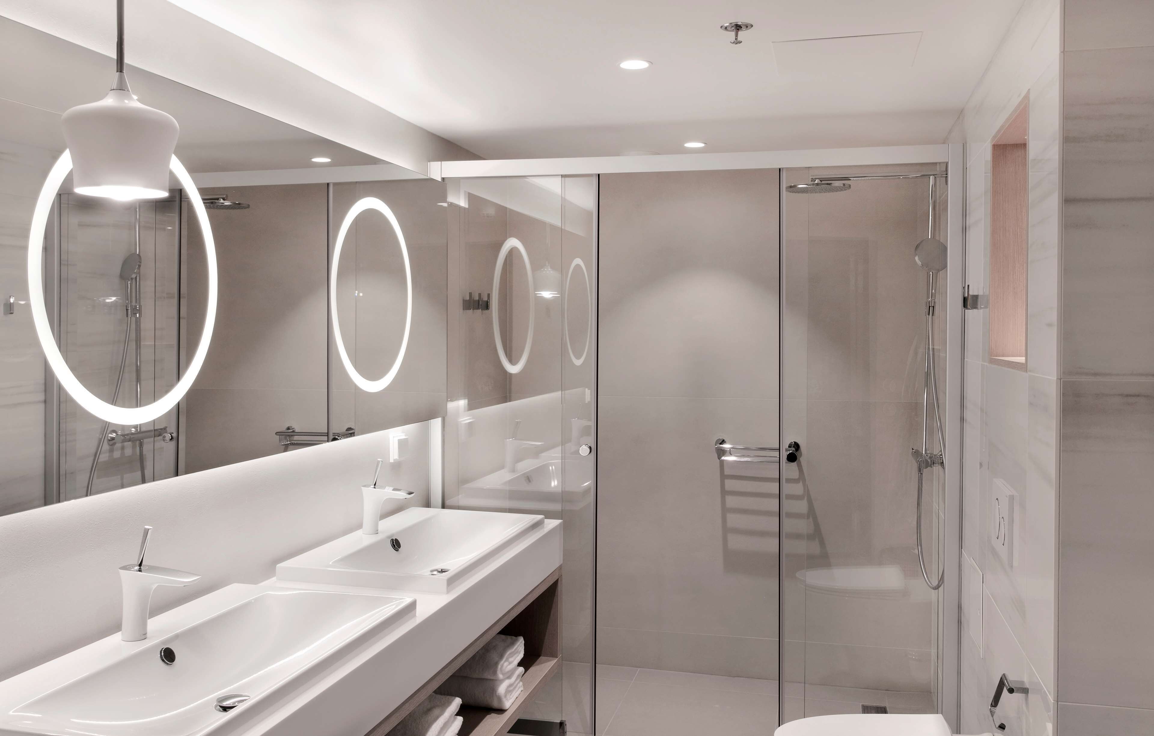 Un baño moderno con un tocador amplio, dos lavamanos, una cabina de ducha de vidrio y un espejo con luminarias circulares.