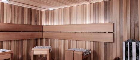 A sauna room with wooden walls and benches, a ceiling light, and a metal rack with stones.