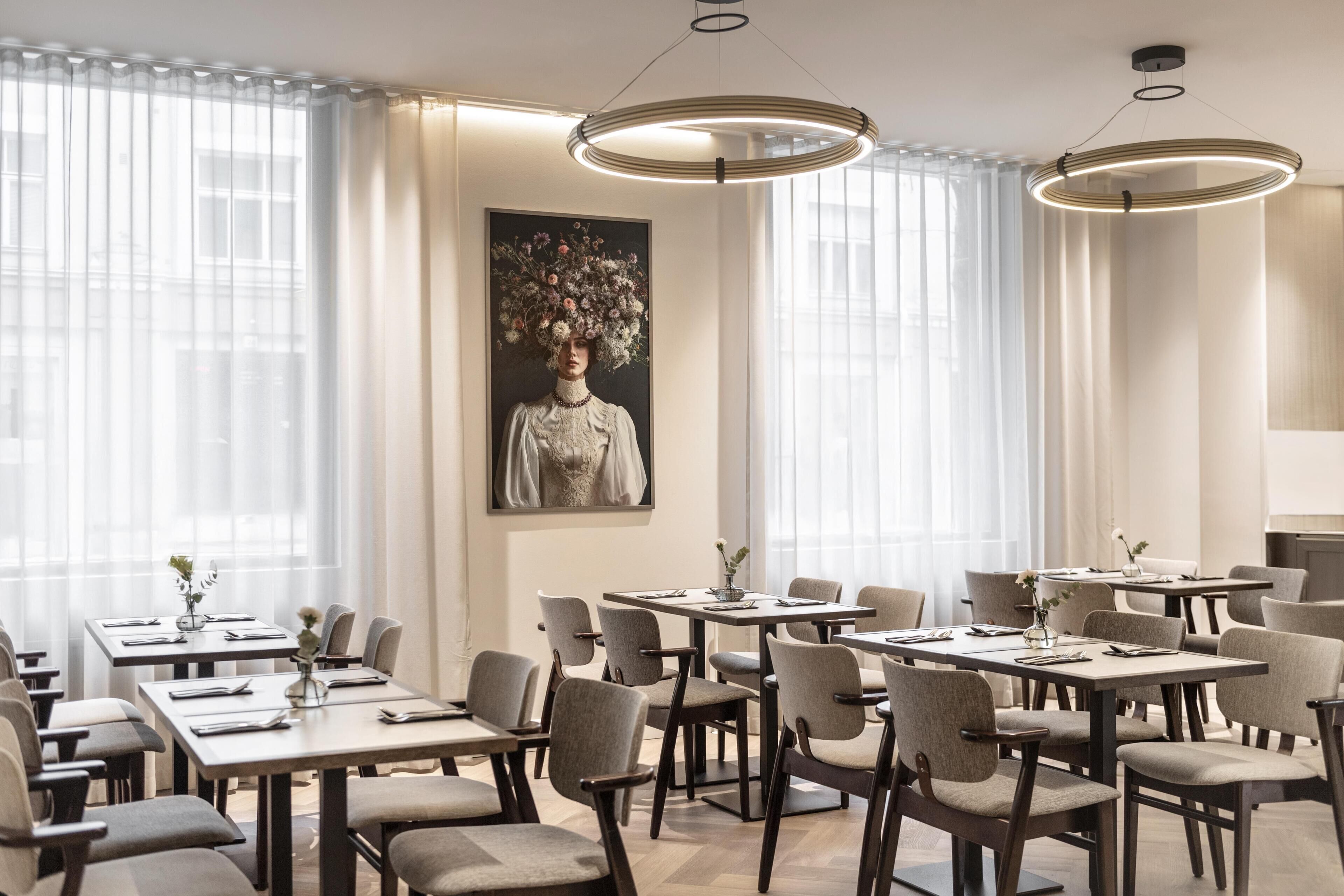 Une salle à manger d’hôtel avec de nombreuses tables et chaises, de grandes fenêtres ornées de rideaux légers et un tableau floral de grande taille accroché au mur.