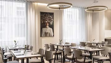 Une salle à manger d’hôtel avec de nombreuses tables et chaises, de grandes fenêtres ornées de rideaux légers et un tableau floral de grande taille accroché au mur.