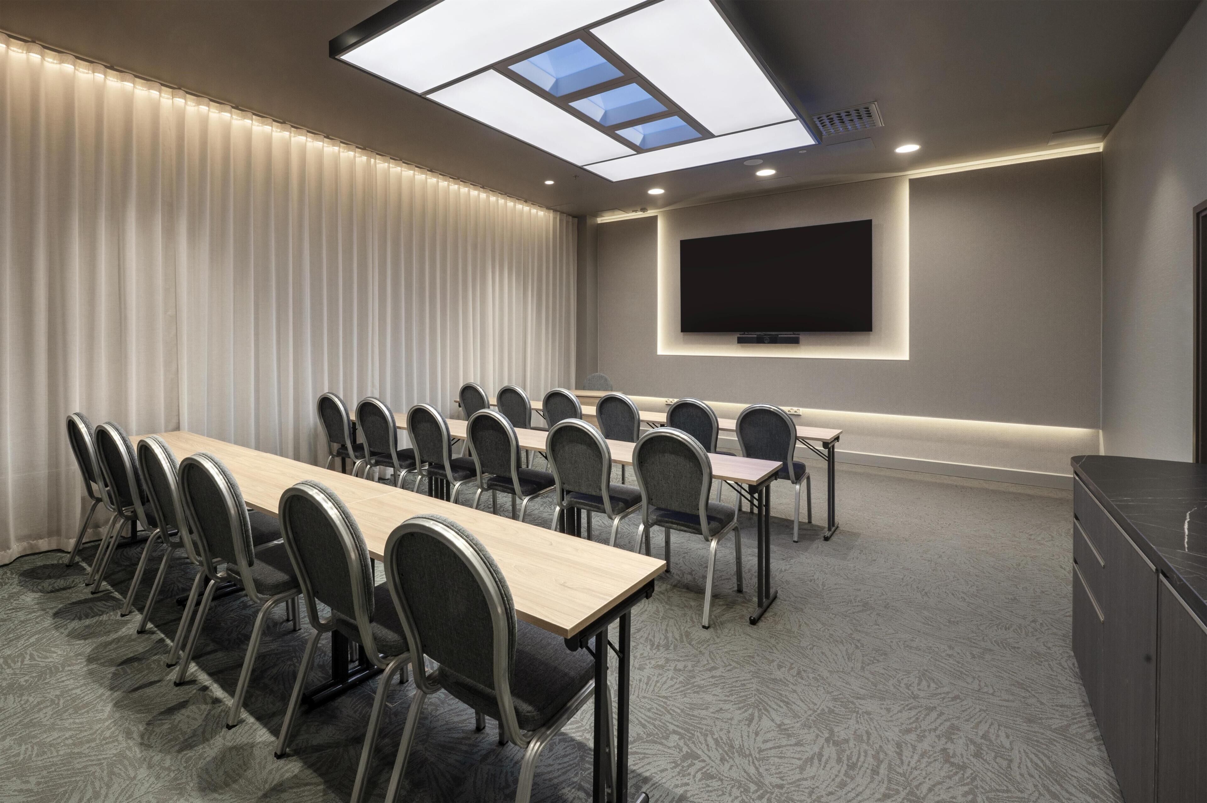 A conference room with a long table, chairs, a flat-screen TV, and a ceiling skylight.