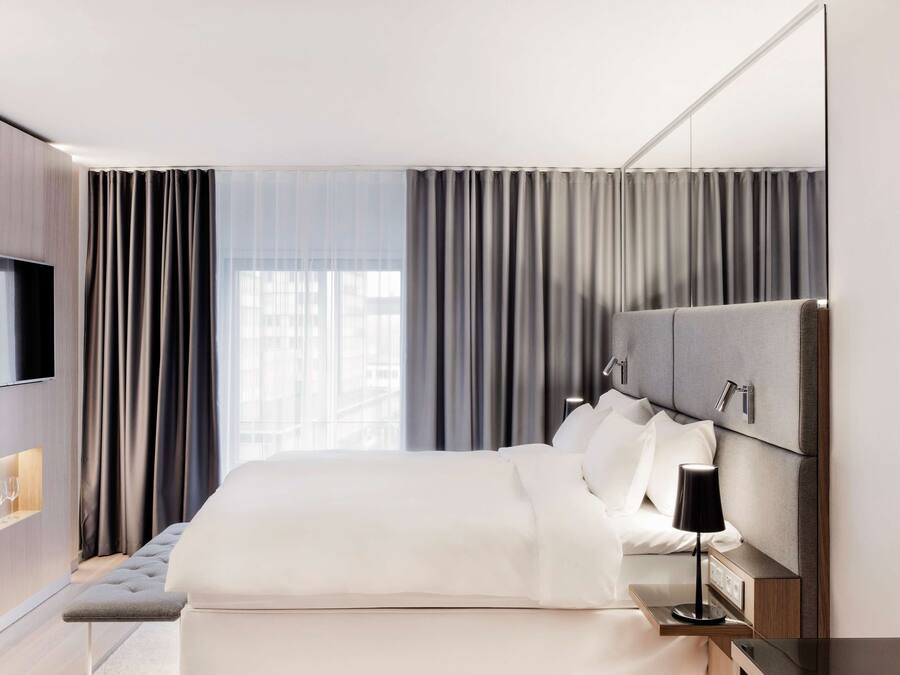 Een moderne hotelkamer met een groot bed, een flatscreen televisie aan de wand, een nachtkastje met een lamp en een groot raam met gordijnen.