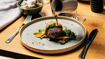 A plate with a steak, vegetables, and sauce, a fork, a knife, and a glass of water on a wooden table.