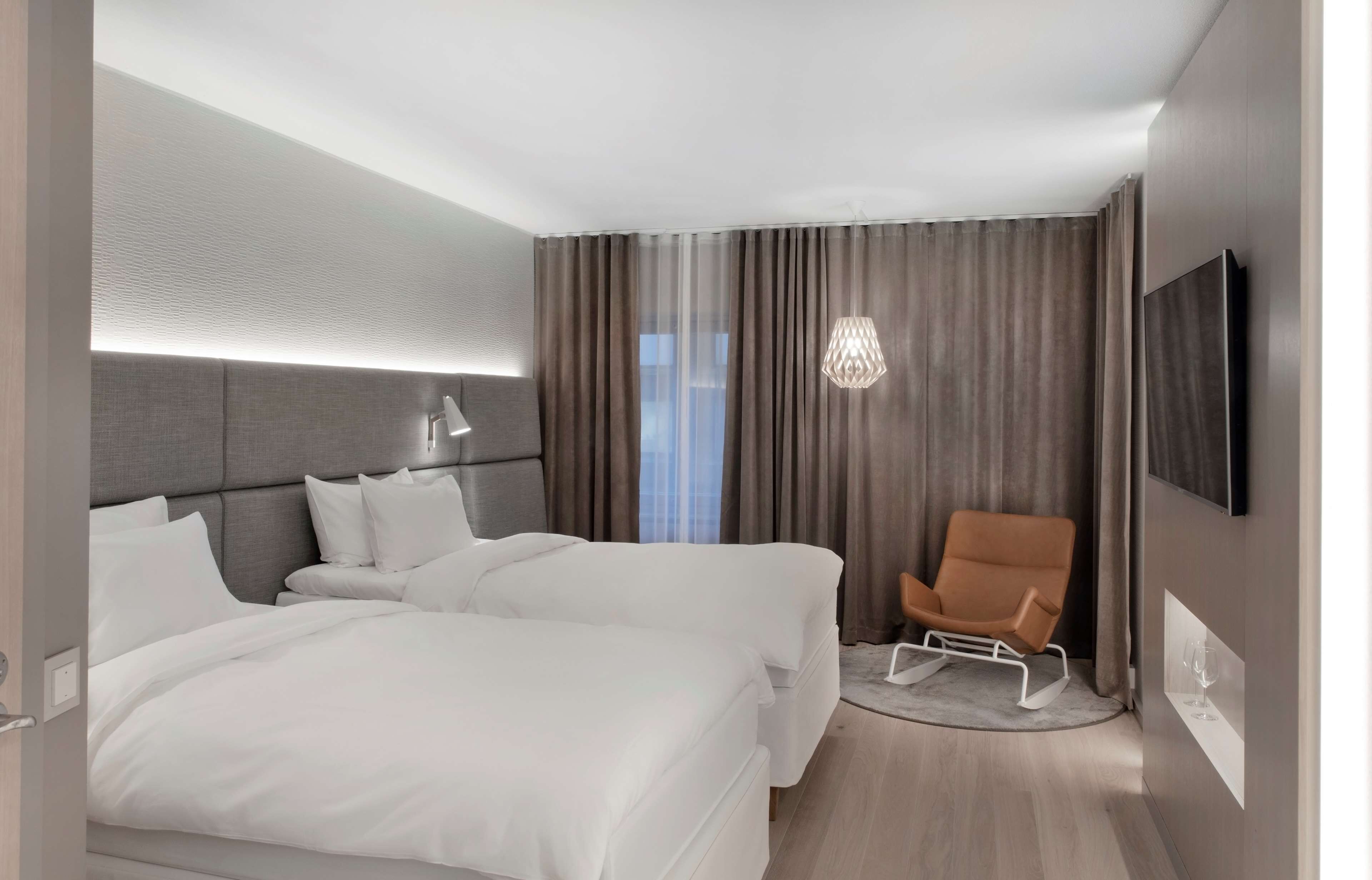 A hotel room with two beds, a chair, a TV, and a window with curtains.
