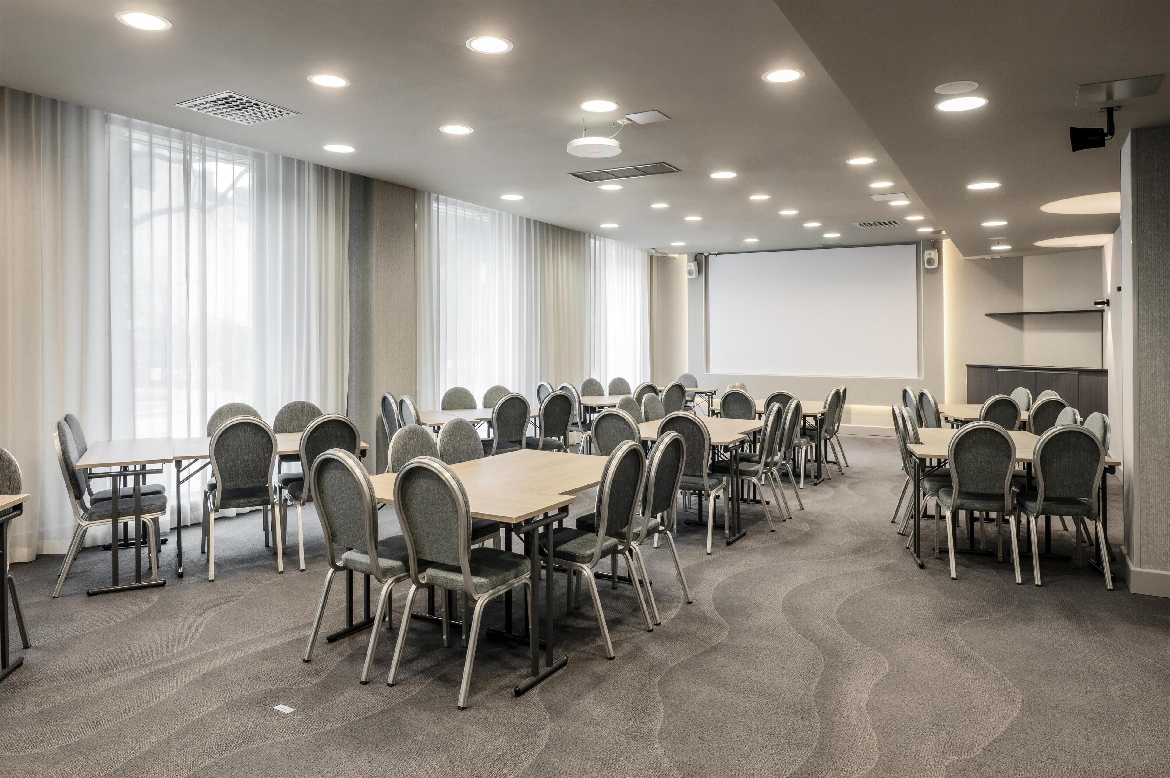 A conference room with multiple tables and chairs, a projector screen, and a large window with curtains.