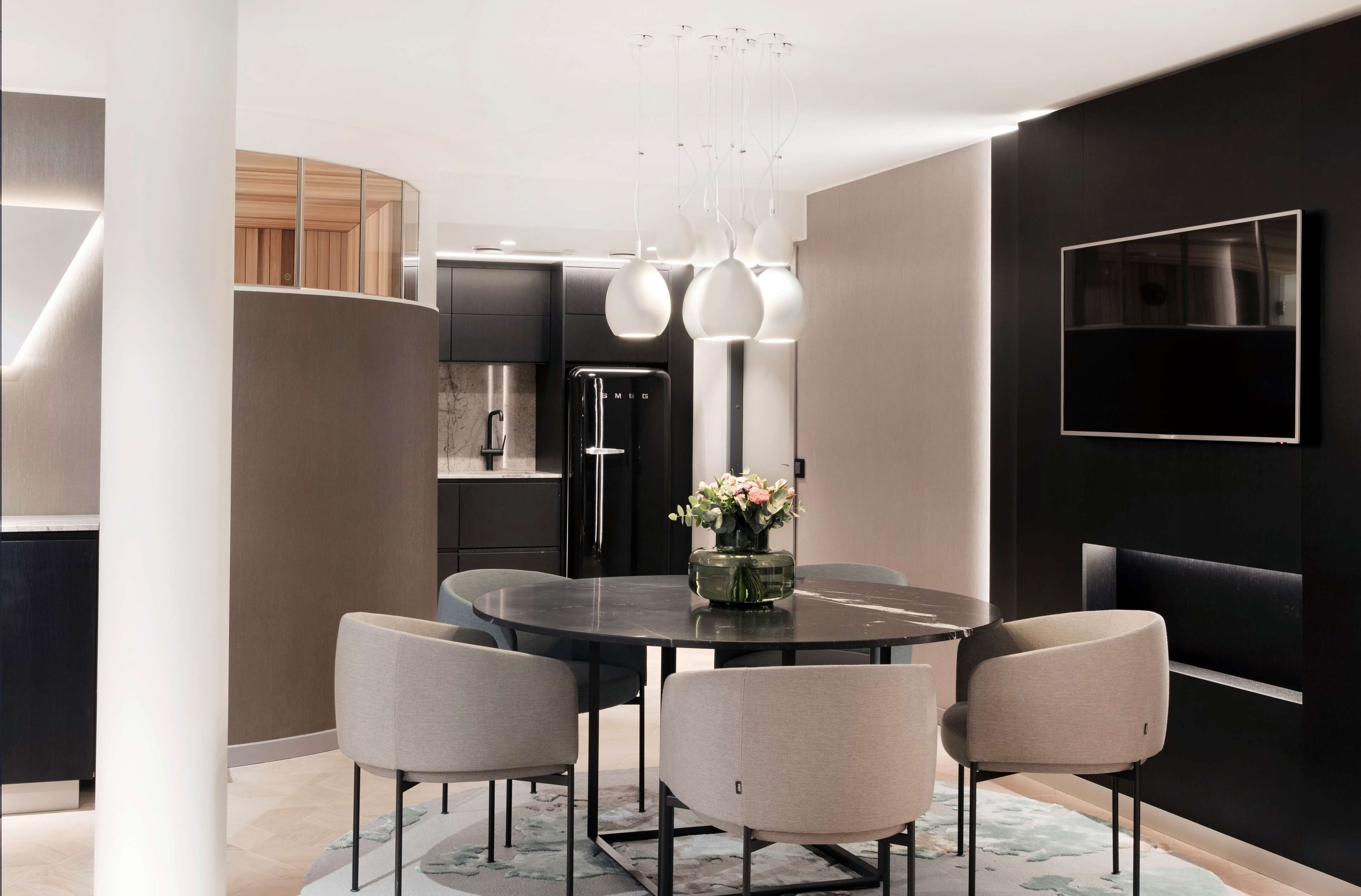 A modern dining area with a round table, four chairs, a vase with flowers, a black refrigerator, and a mounted TV on the wall.