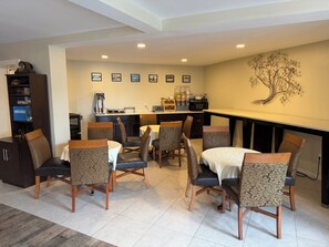 A dining area with a kitchen in the background, featuring a coffee machine, microwave, and various kitchen appliances. - Governor House Inn (Falls Church)