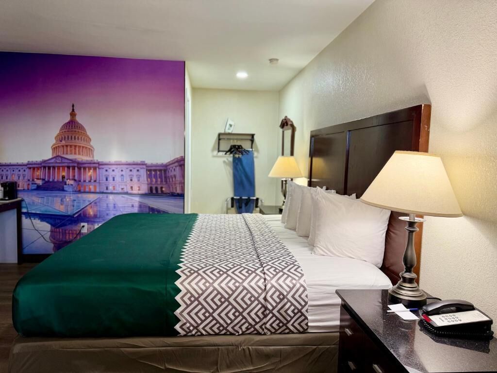 Une chambre d’hôtel avec un lit, un bureau doté d’une lampe, un téléphone, un tableau représentant le Capitole et une table de chevet.