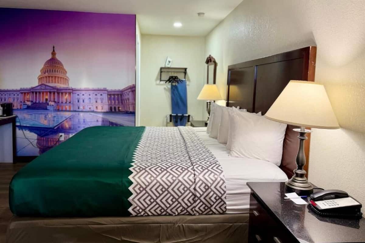 A hotel room with a bed, a desk with a lamp, a phone, a painting of the Capitol building, and a nightstand.