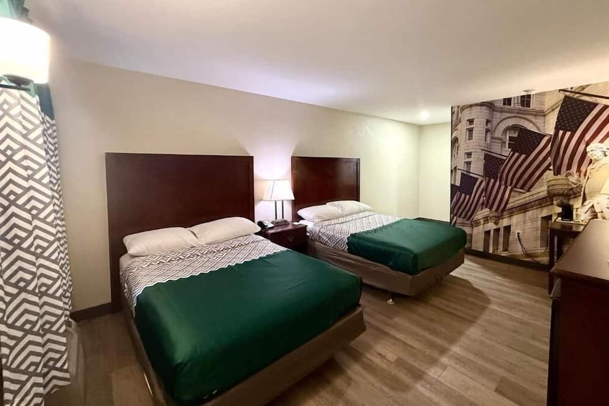 A hotel room with two beds, a nightstand, a lamp, and a large American flag mural on the wall.