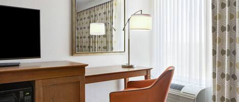 Habitación de hotel con un escritorio de madera, televisor, silla, lámpara y un ventanal con cortinas.
