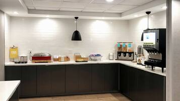 A modern kitchen with white tiled walls, a black pendant light, a coffee machine, and various food items on the counter.