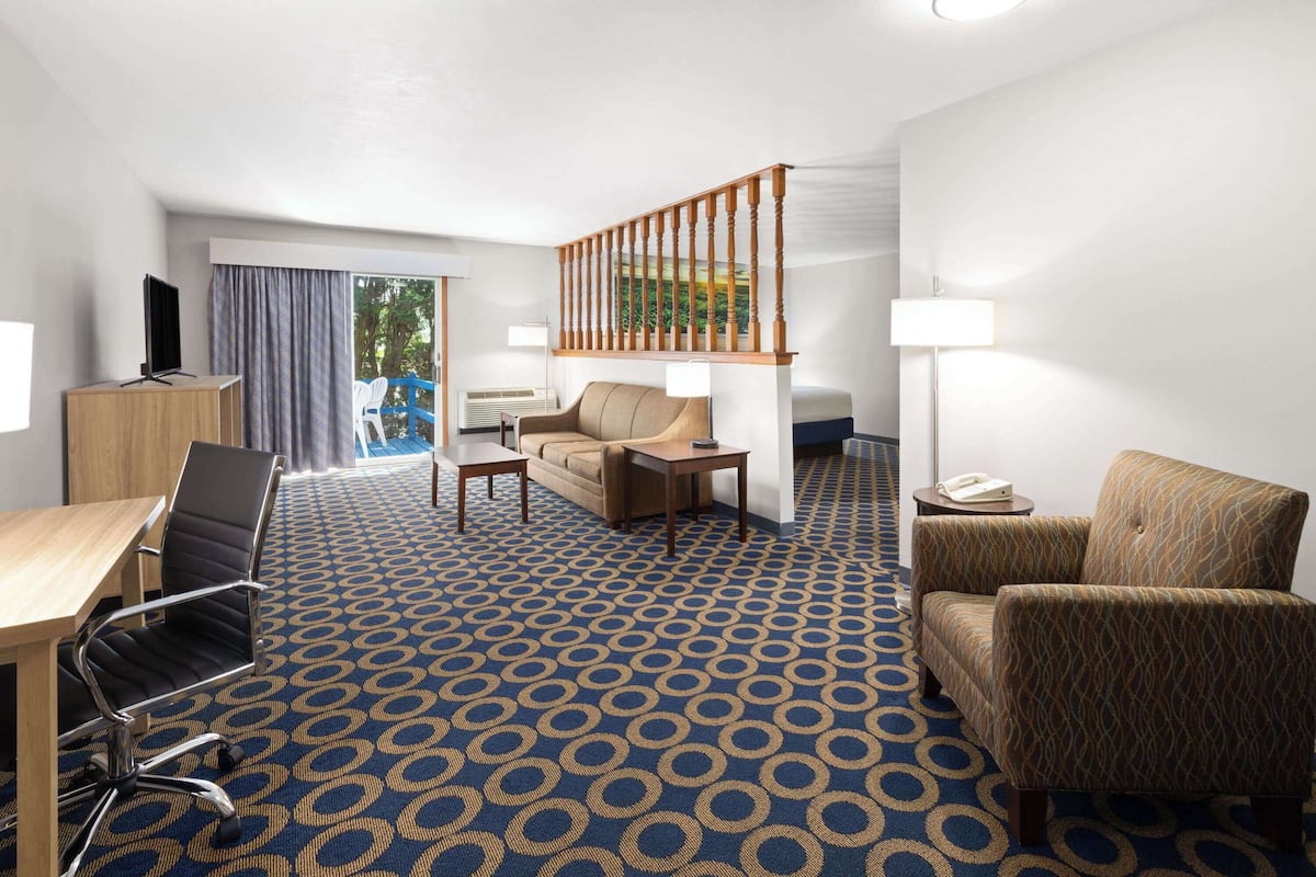 A hotel room with a blue patterned carpet, a wooden desk with a chair, a sofa, a small table, a lamp, and a staircase with a railing.