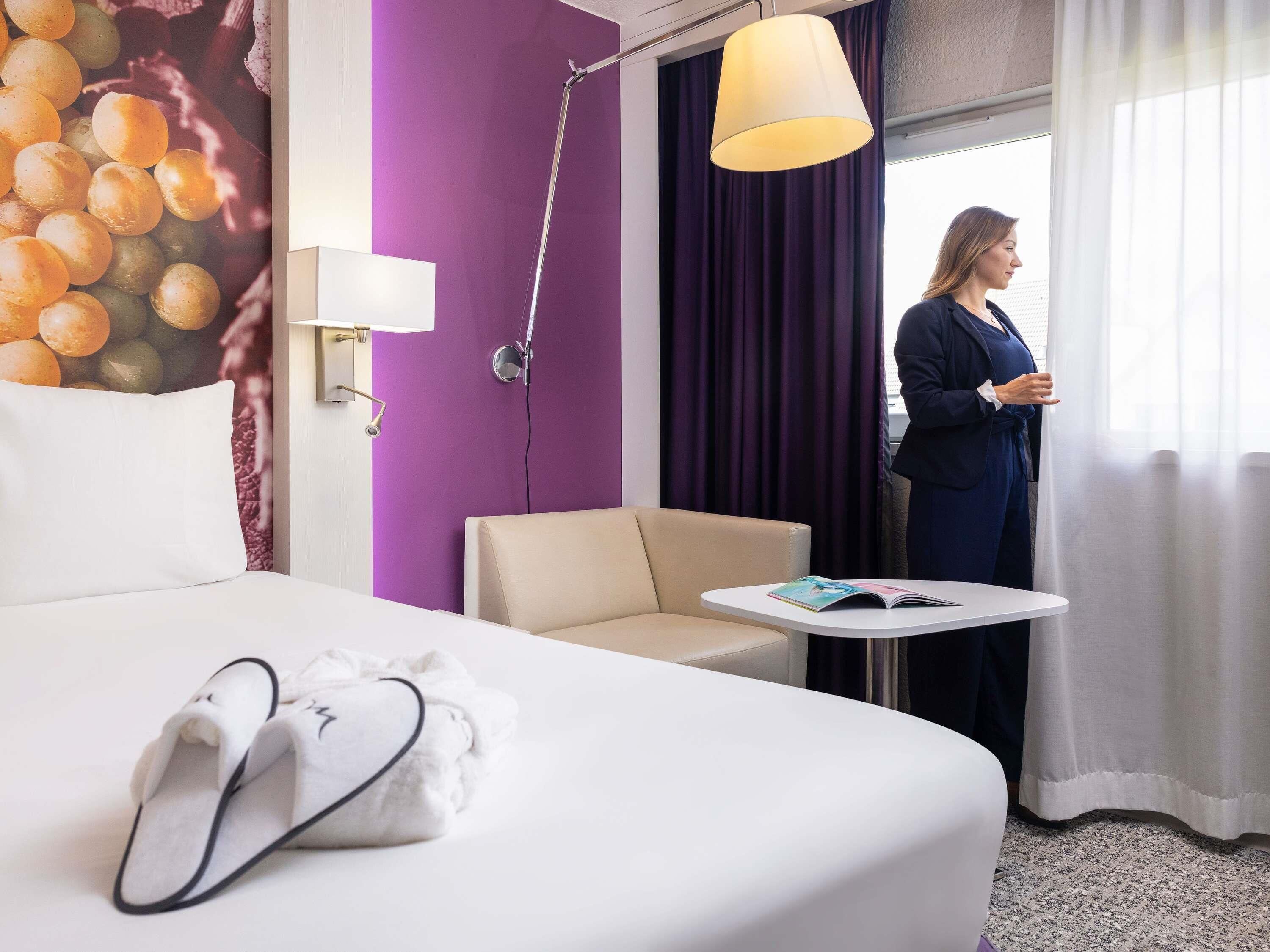 A hotel room with a bed, a small table, a chair, a lamp, and a window with curtains.