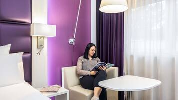Een moderne hotelkamer met een paarse accentmuur, een bed, een zithoek met een tafel en een groot raam met gordijnen.