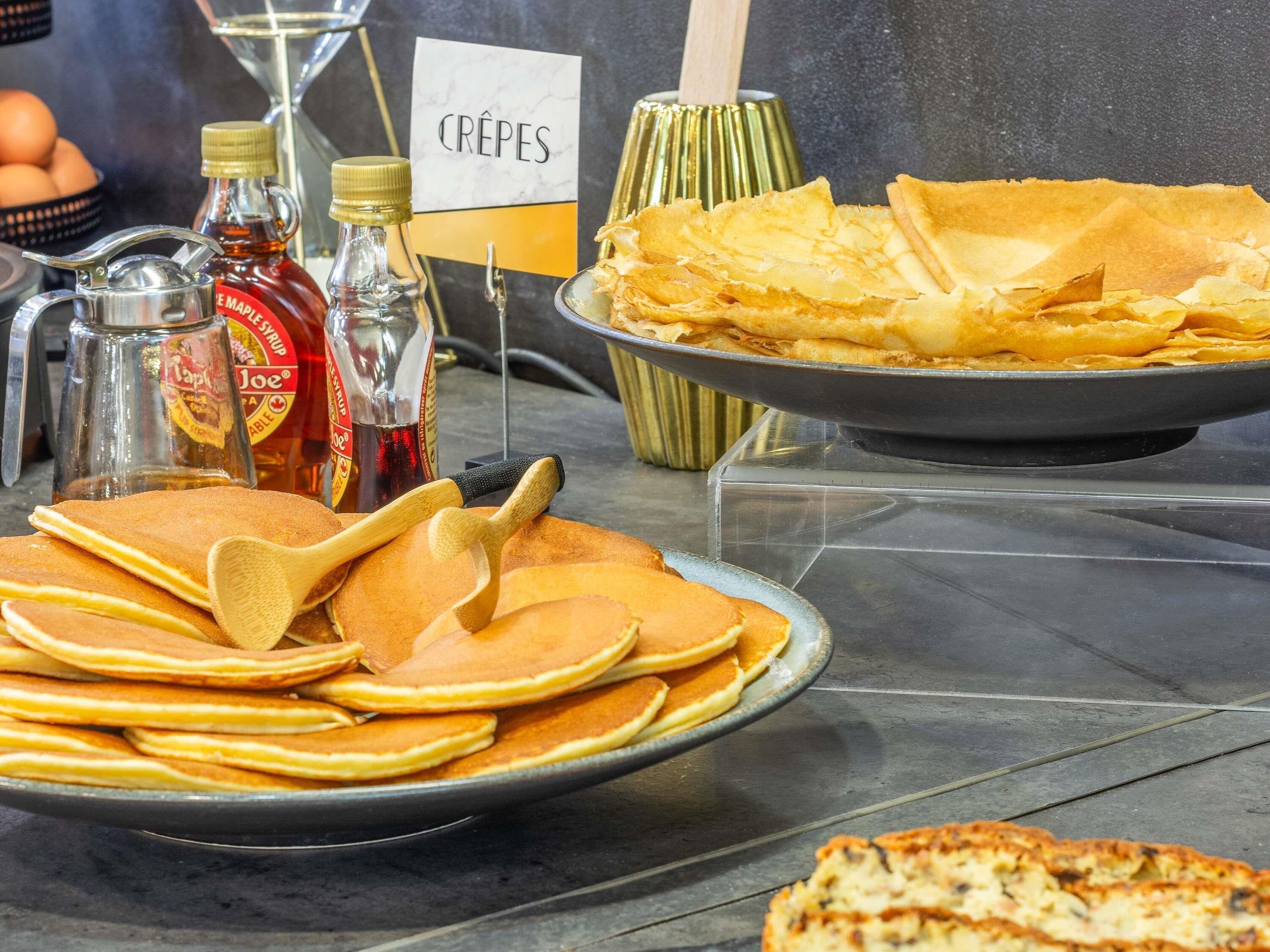 Un buffet de petit-déjeuner proposant des crêpes, des pancakes, du sirop, et un panneau indiquant l’emplacement des crêpes.