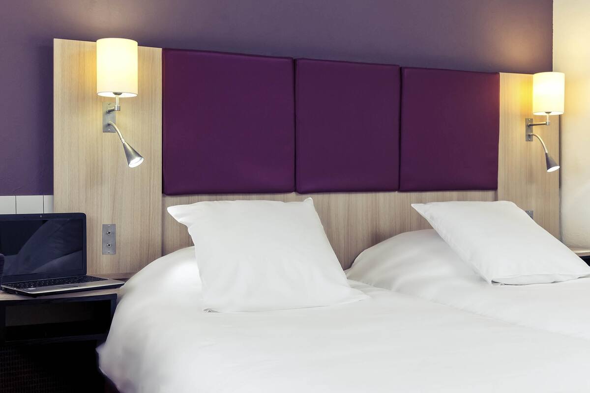 Ein Hotelzimmer mit einem Bett, zwei Nachttischlampen, einem Laptop auf einem Nachttisch und einem hölzernen Kopfteil mit violetten Paneelen.