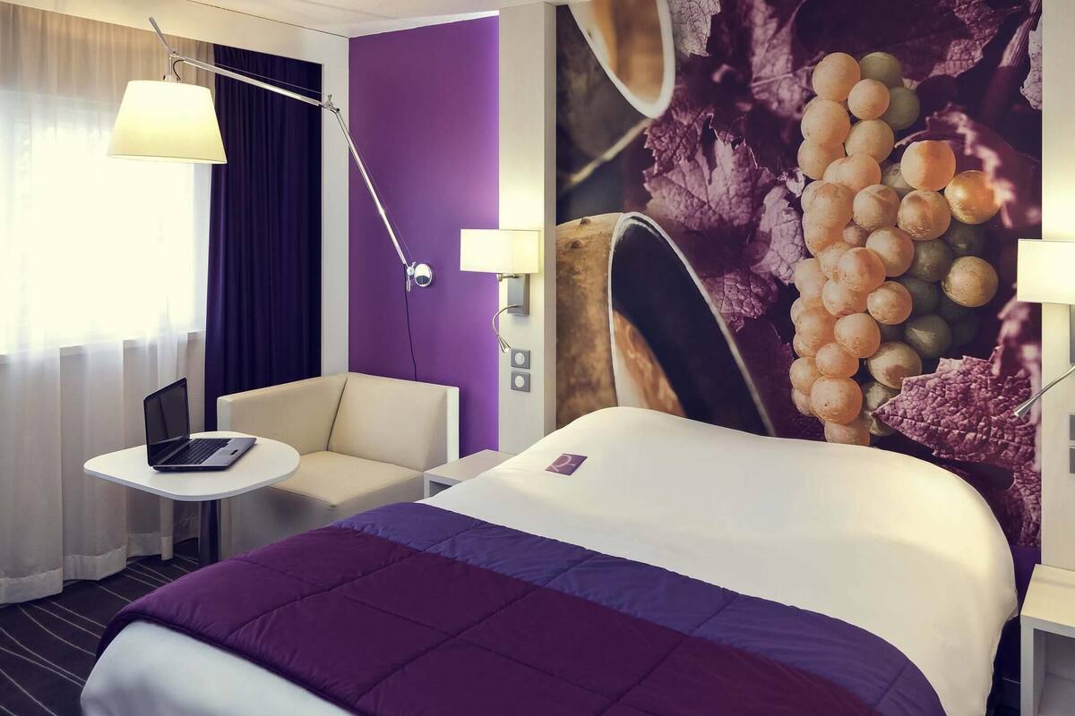 Ein Hotelzimmer mit Bett, Schreibtisch, Stuhl, Lampe und Wandverzierungen mit Traubenmotiv.