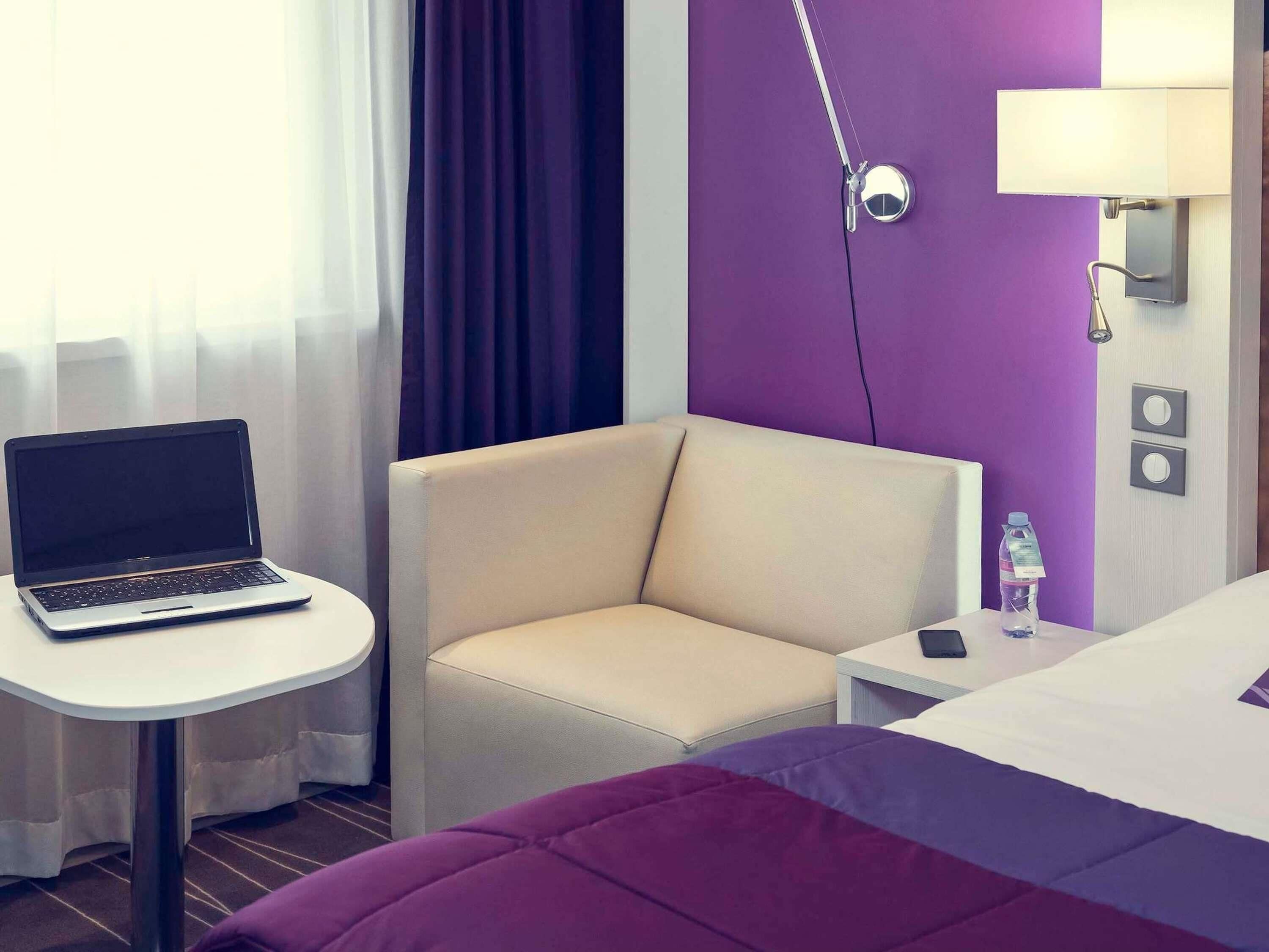 Et hotelværelse med en bærbar computer på et lille bord, en beige lænestol, en seng med et lilla og hvidt sengetæppe og en vandflaske på sengebordet.