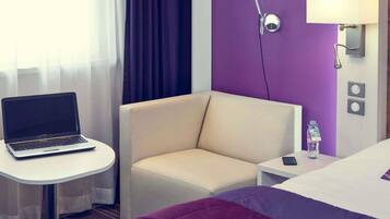 Et hotelværelse med en bærbar computer på et lille bord, en beige lænestol, en seng med et lilla og hvidt sengetæppe og en vandflaske på sengebordet.