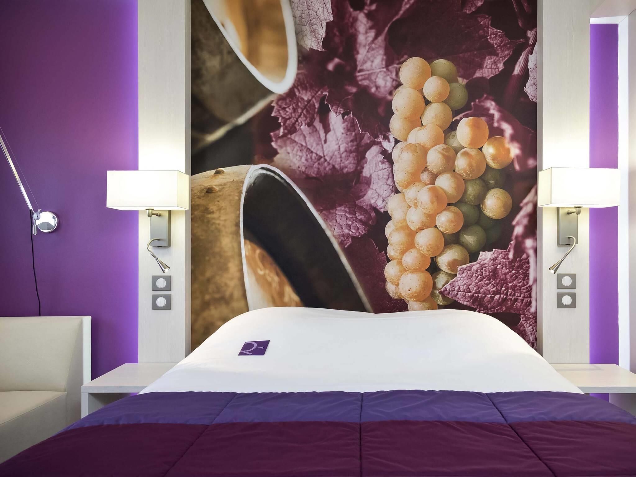 A hotel room with a bed, a wall-mounted TV, and a nightstand with a lamp.