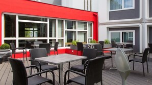 Een terras buiten met meerdere tafels en stoelen, grote ramen en een gevel van een gebouw in rood en wit.