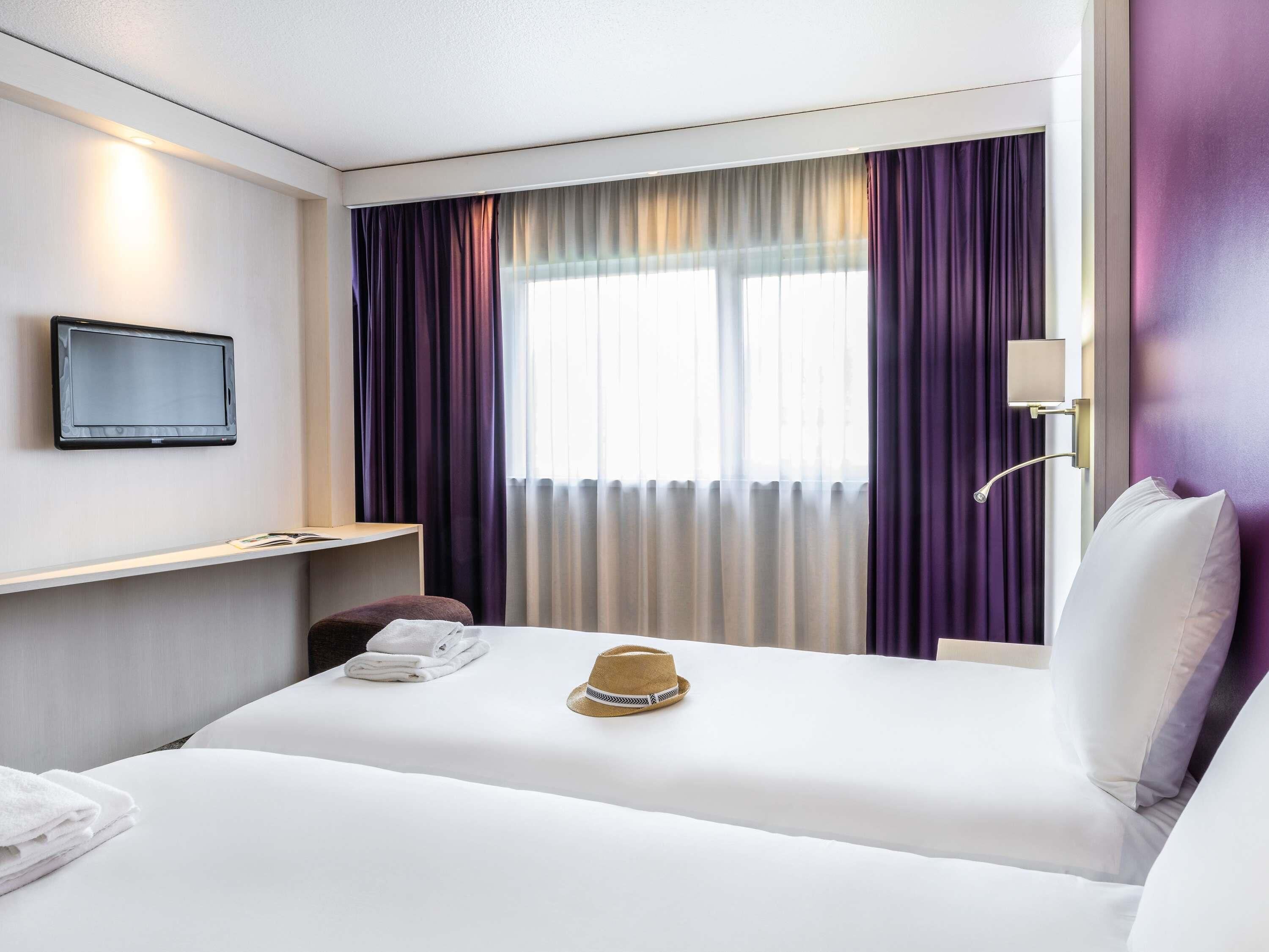 Habitación de hotel con cama, televisor montado en la pared, mesita de noche con lámpara y ventana con cortinas.