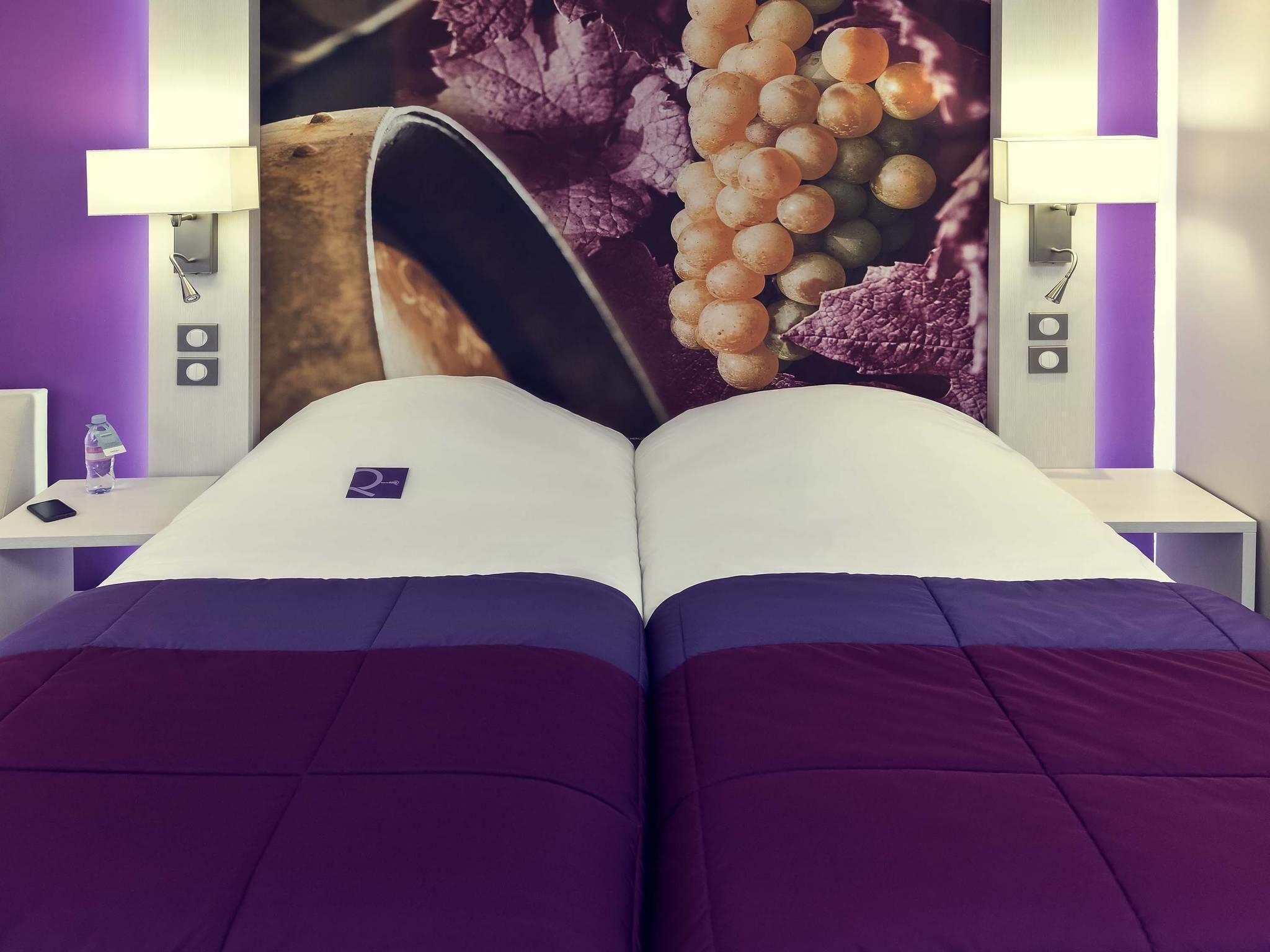 Una cama doble con una colcha morada y blanca, una mesita de noche con una botella de agua y una lámpara fijada a la pared.