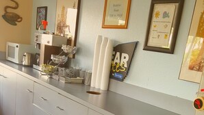 Una cocina con microondas, tostadora y cafetera. Hay cuadros en la pared y un letrero que indica el horario del desayuno.