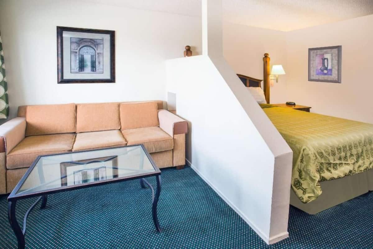 A hotel room with a sofa, a glass-top table, a bed with a green bedspread, a nightstand with a lamp, and two framed pictures on the walls.