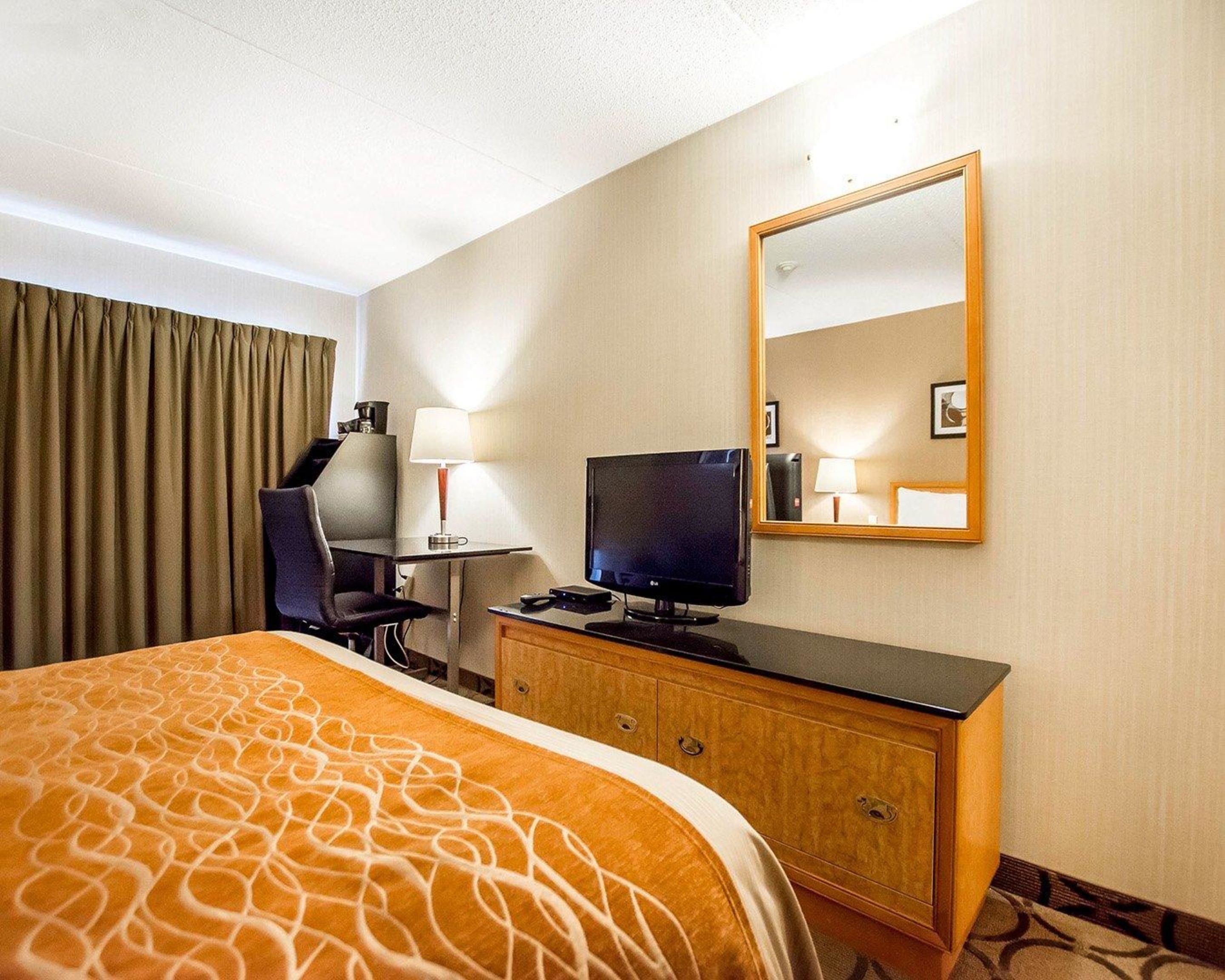 Habitación de hotel con una cama, un escritorio con computadora, una silla, un televisor, un aparador y un espejo.
