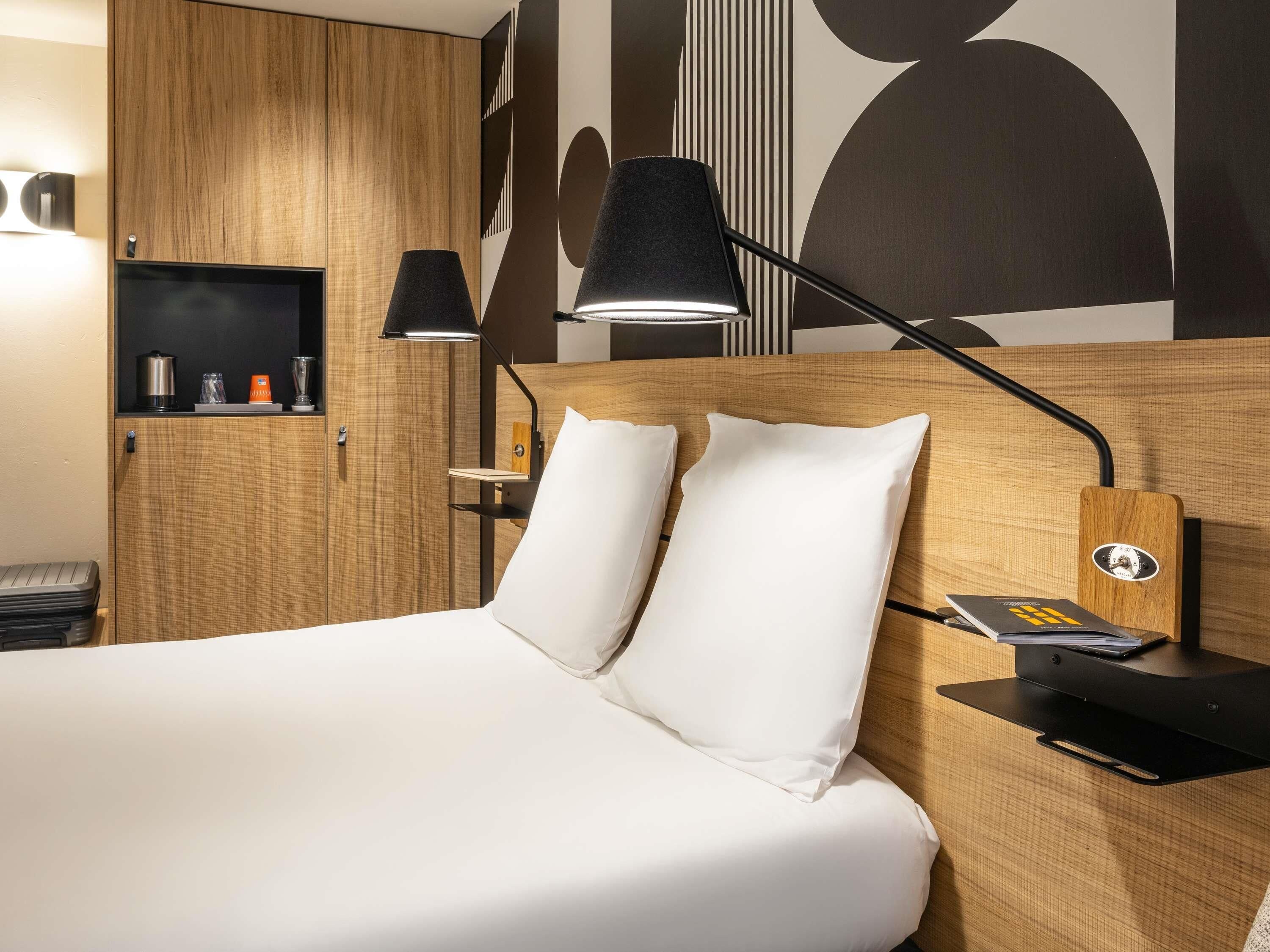 A hotel room with a wooden headboard, a bed with white linens, a nightstand with a lamp, and a shelf with toiletries.