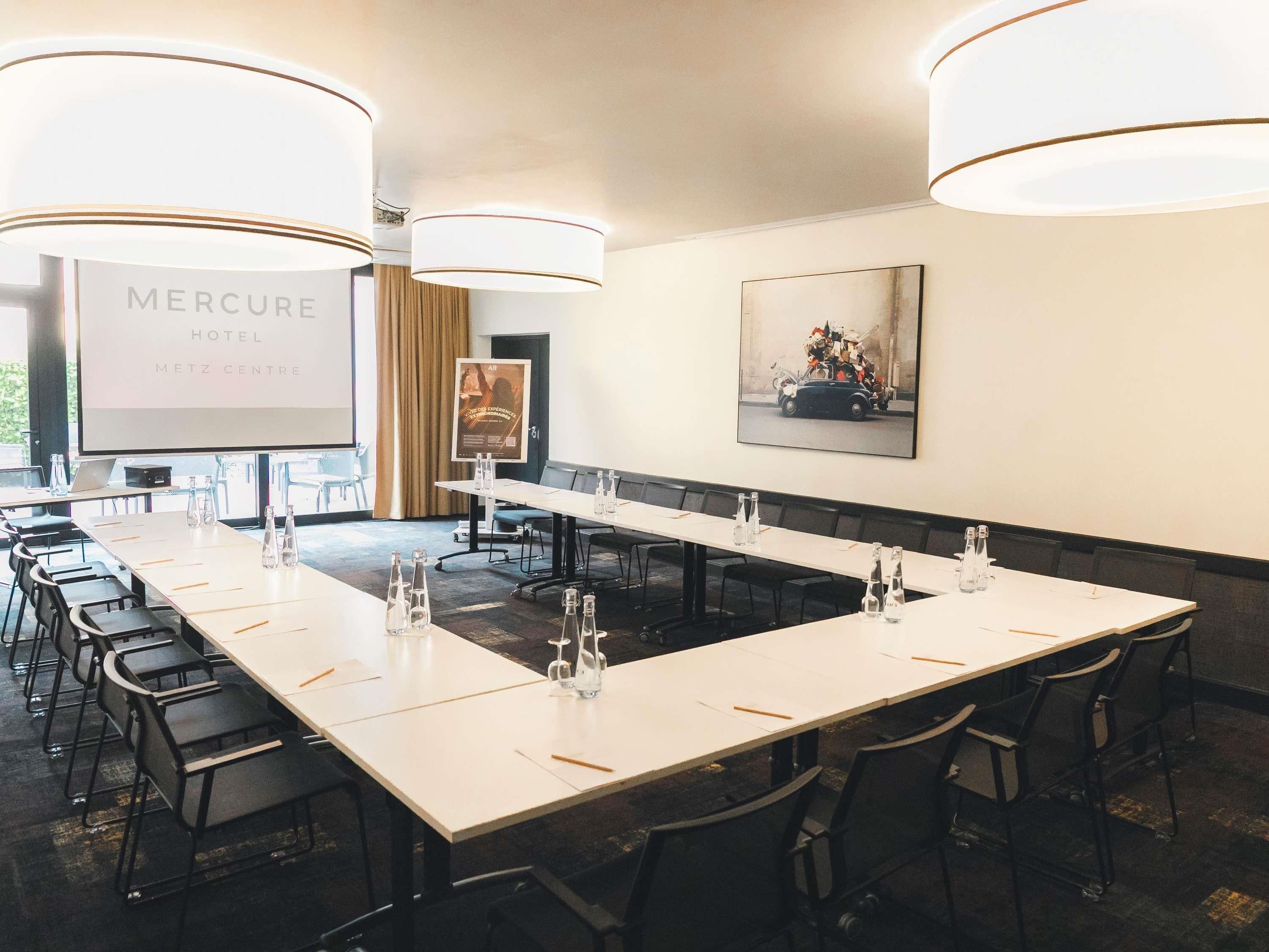 Une salle de conférence équipée d’une longue table, de chaises et d’un écran de projection affichant le texte « Hôtel Mercure Metz Centre ».