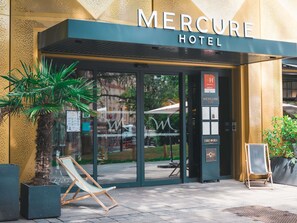 The exterior of the Mercure Hotel with a glass entrance, a palm tree, and a chair outside.