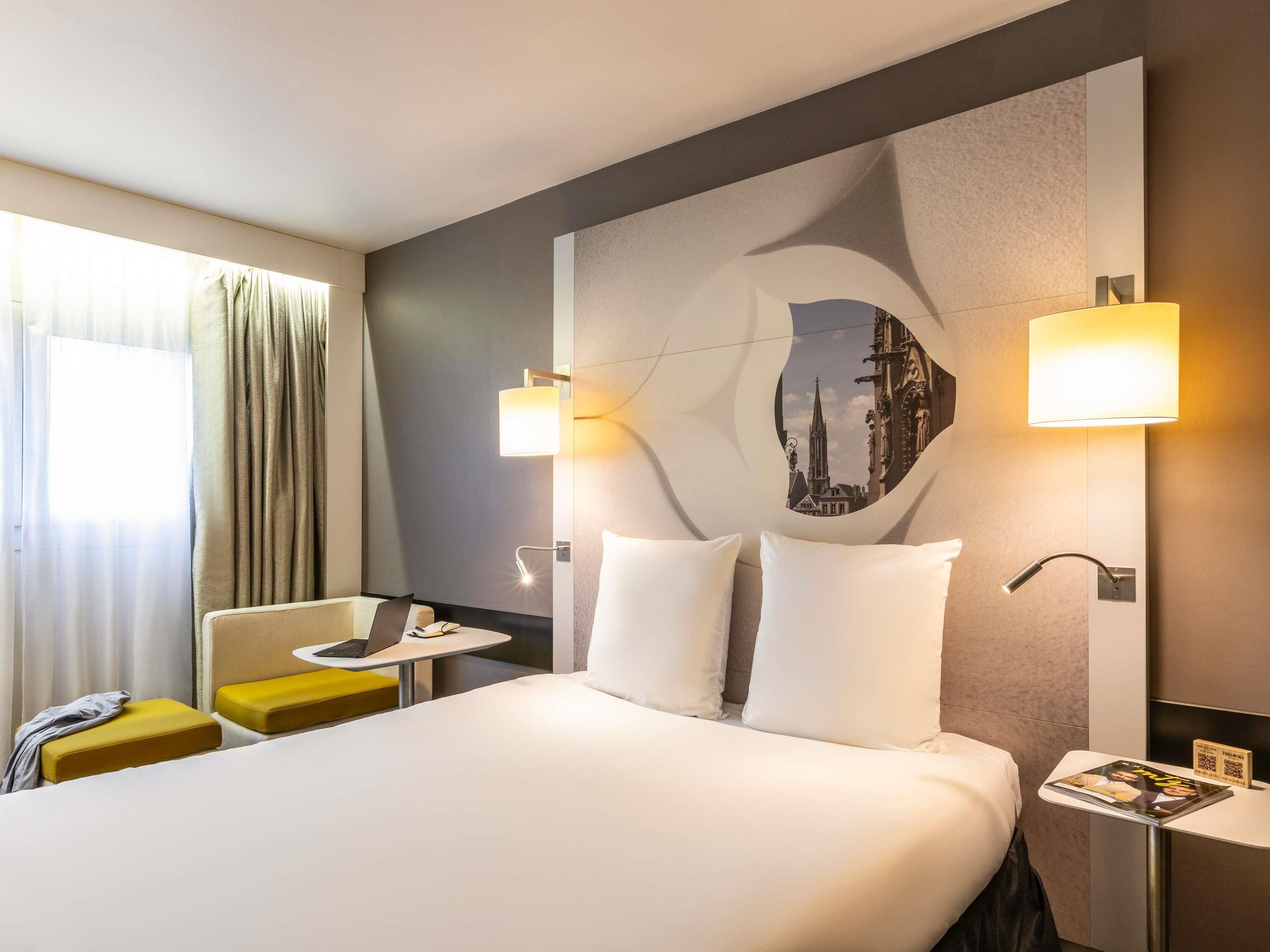 A hotel room with a bed, a desk, a chair, and a wall-mounted lamp.