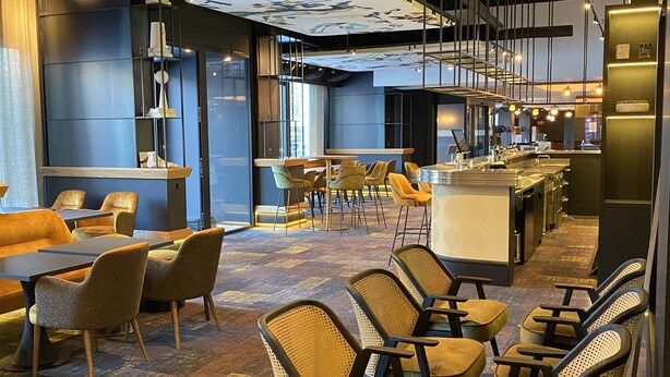 Un interior moderno con zona de bar, asientos con sillas amarillas y marrones, y un techo estampado.