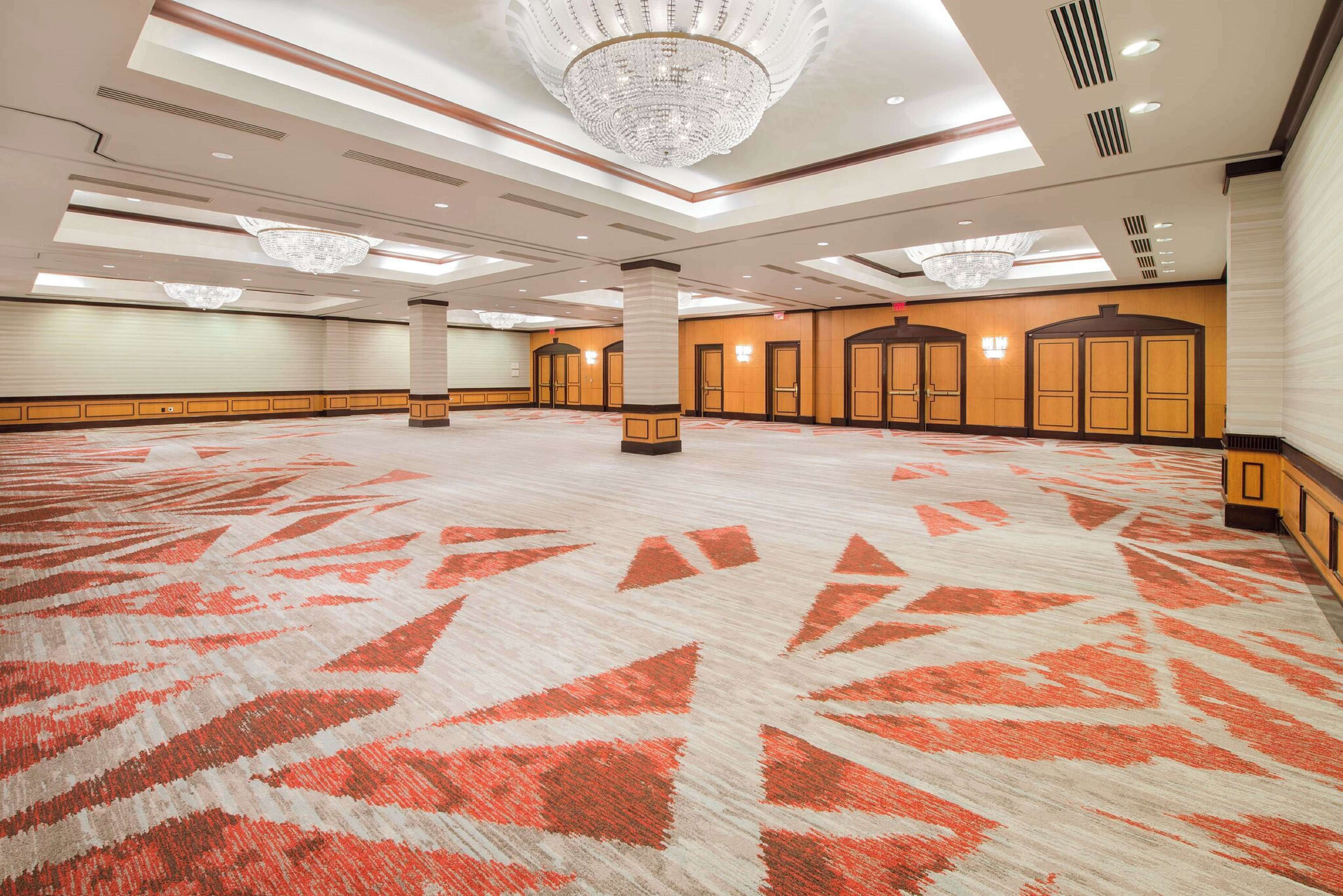 A spacious hotel lobby with a patterned carpet, large chandeliers, and a row of doors.