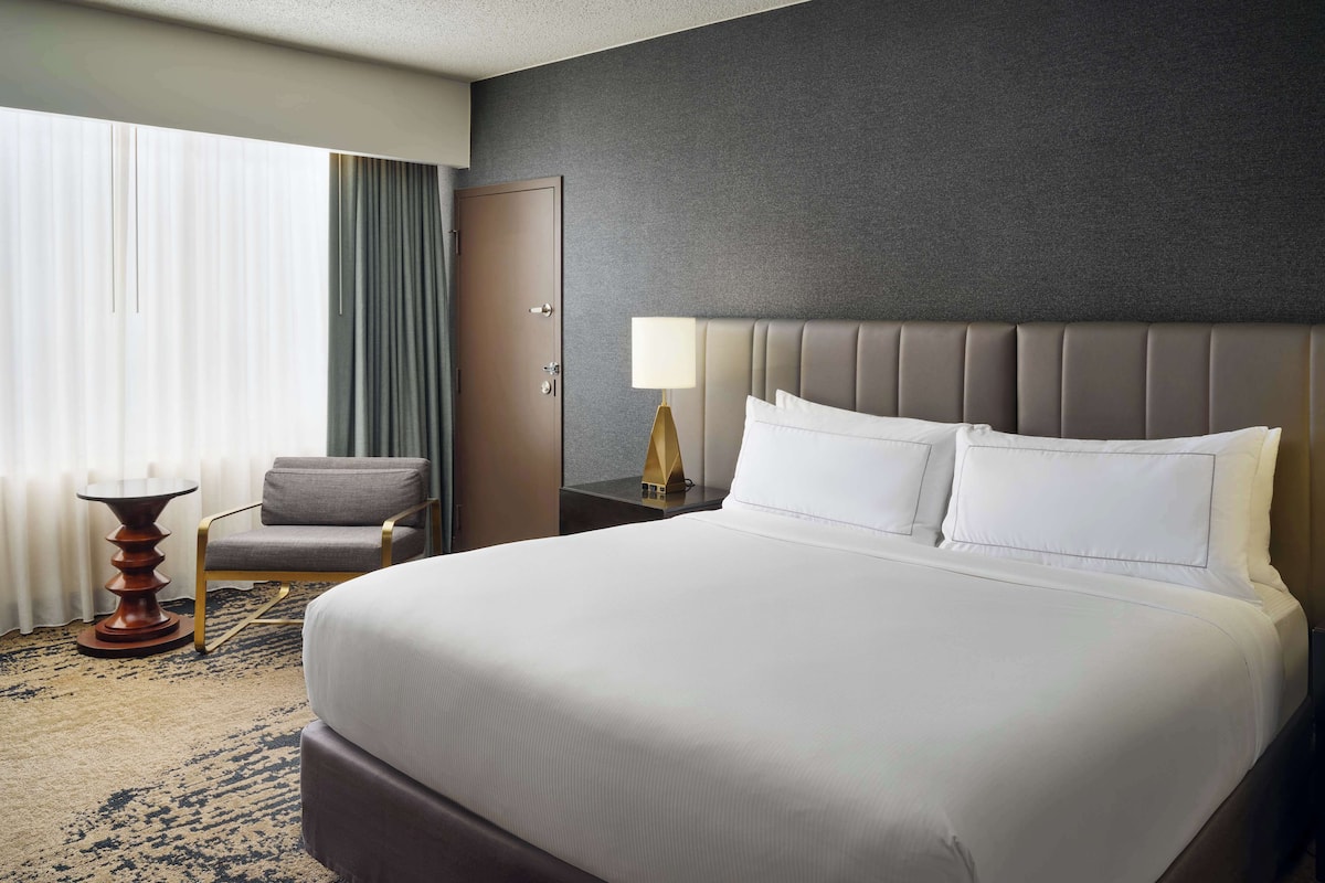 A hotel room with a large bed, a chair, a nightstand, a lamp, and a window with curtains.