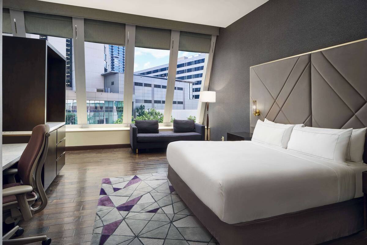 A hotel room with a large bed, a desk with a chair, a sofa, and a view of a building through the window.