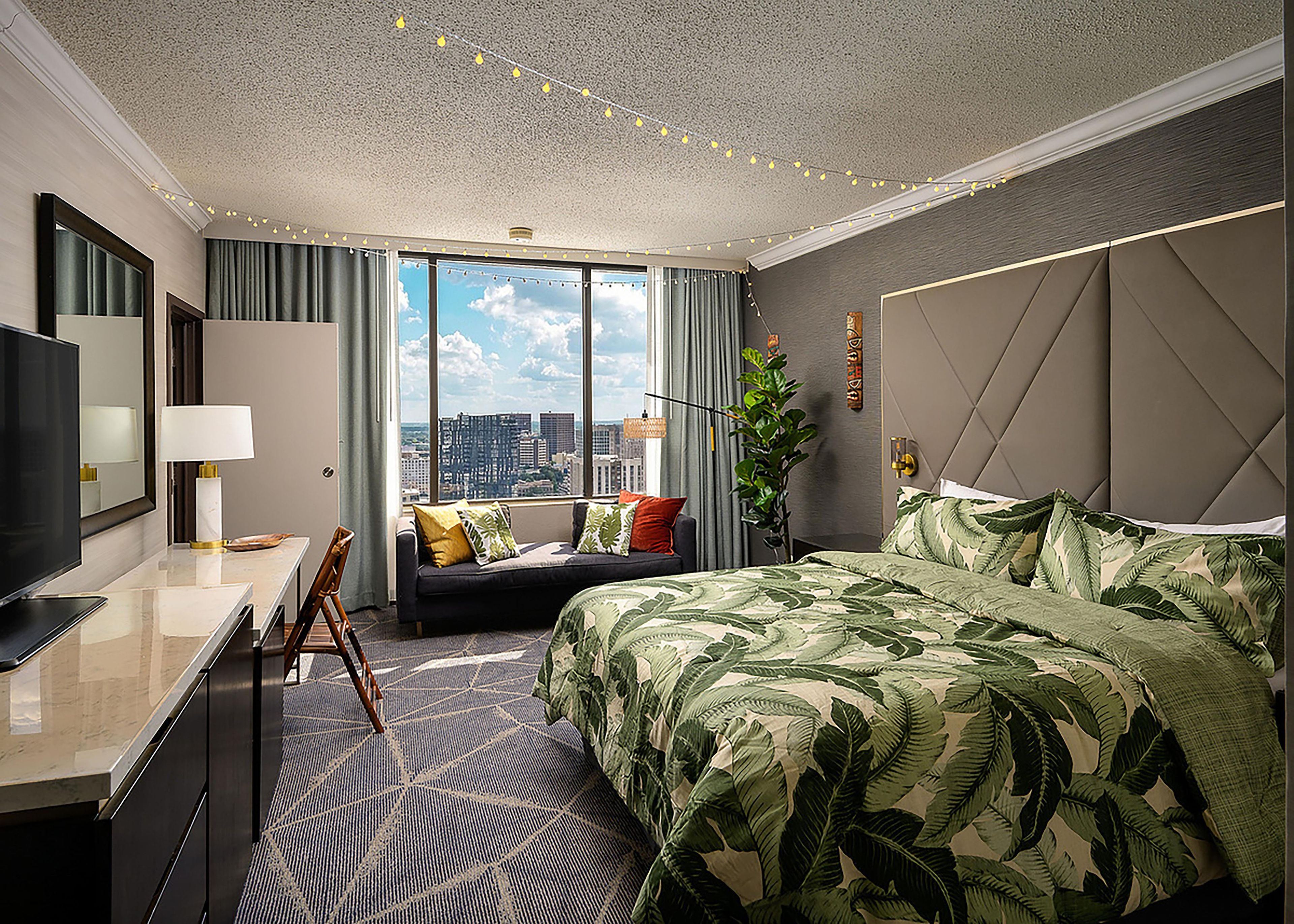 A hotel room with a large bed, a desk with a TV, a sofa, and a view of the city.