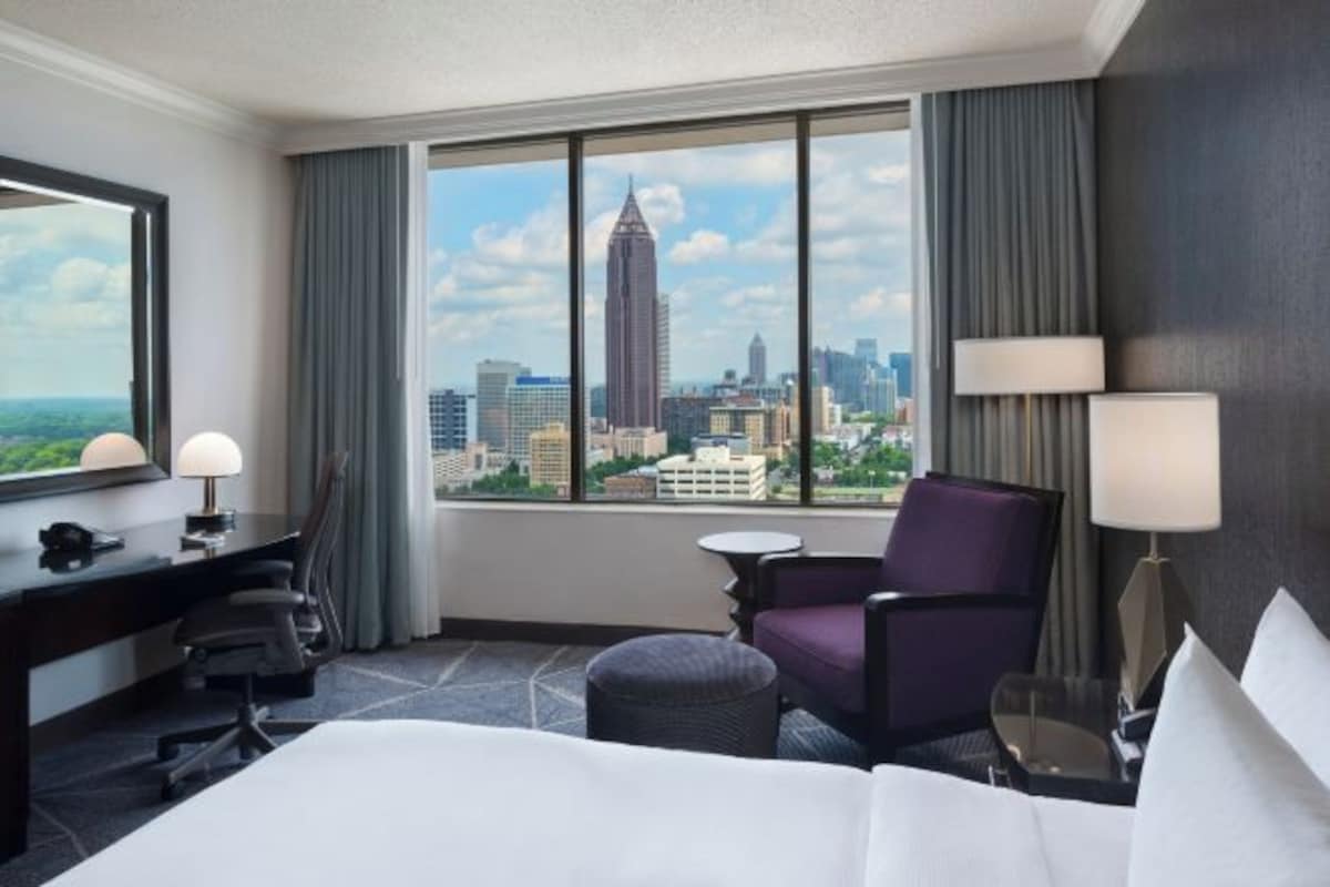 A hotel room with a large window offering a city view, a bed, a desk with a chair, a small table, and a purple armchair.