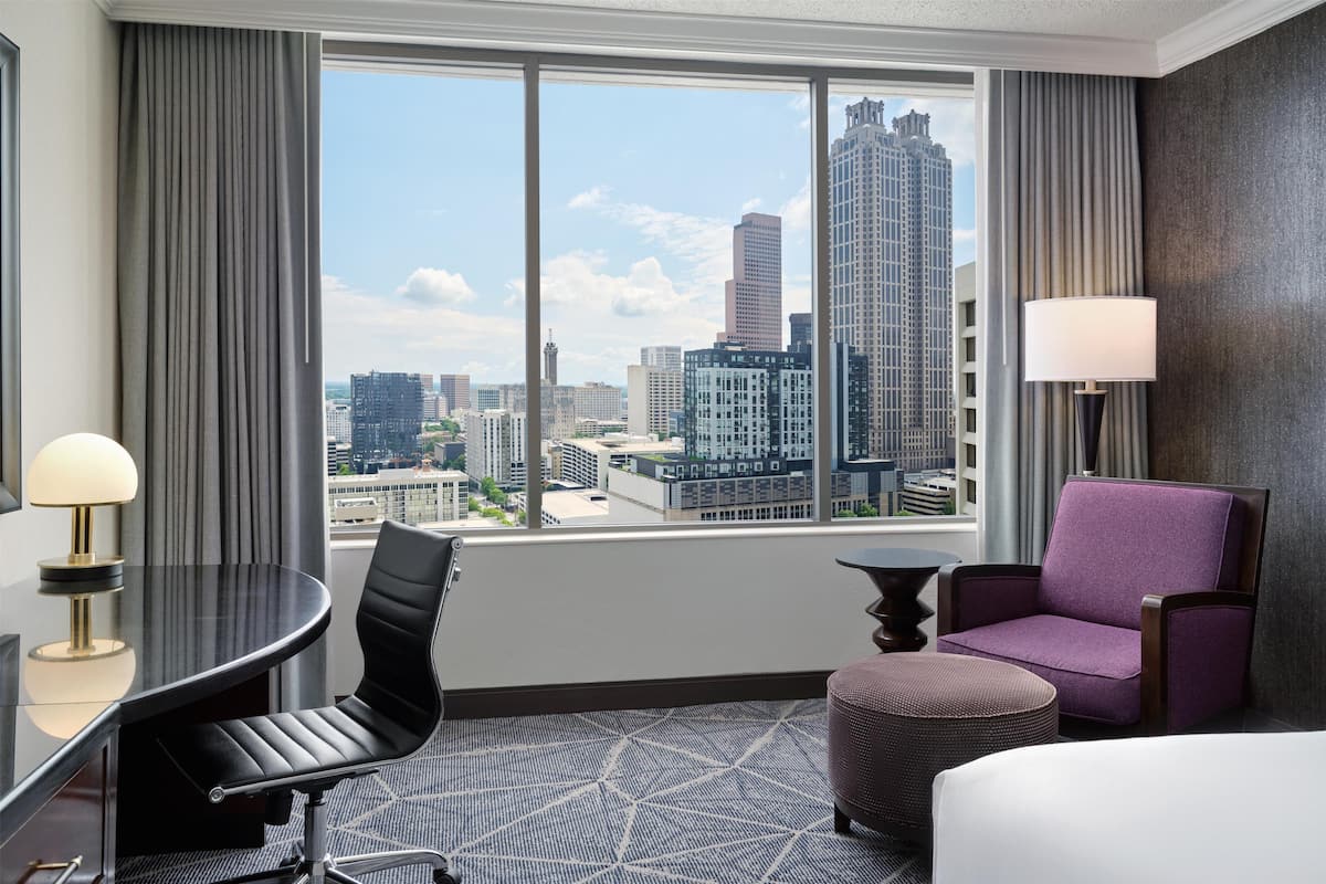 A hotel room with a city view, a round table, a purple armchair, and a small ottoman.
