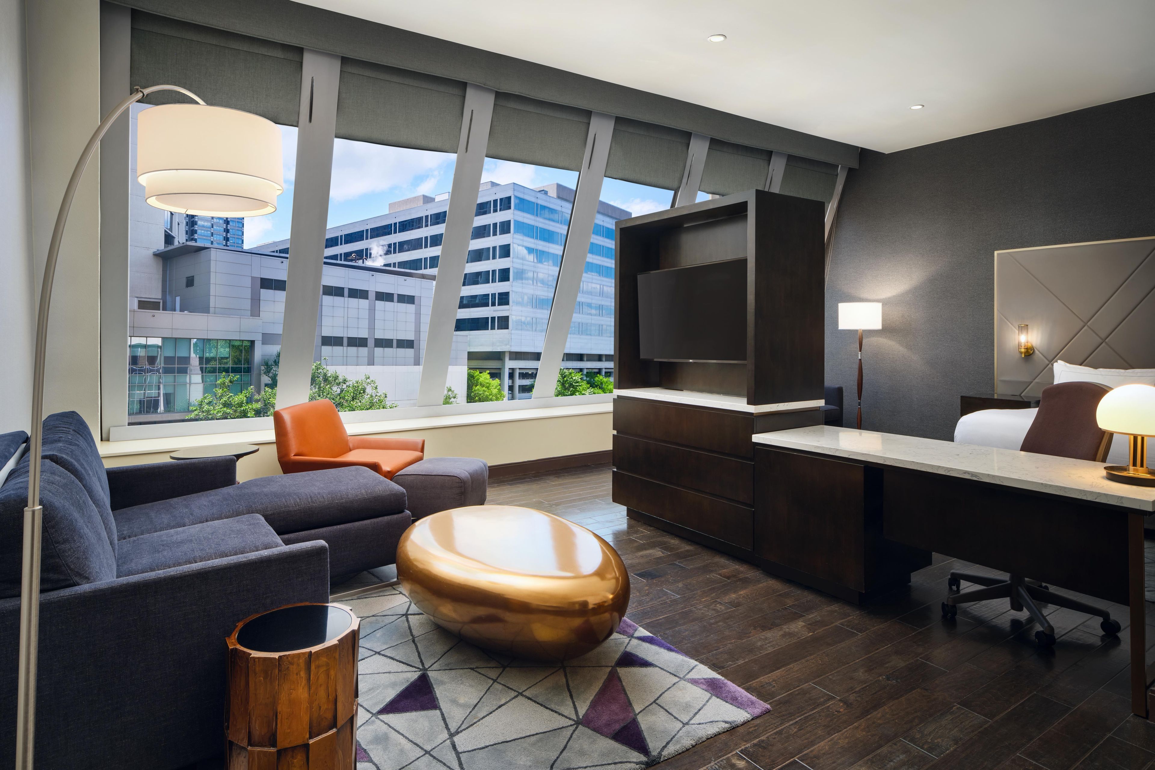 A modern hotel room with a sofa, a round wooden coffee table, a desk with a lamp, and a large window offering a city view.