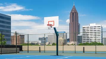 Una cancha de baloncesto al aire libre con aro, rodeada por una cerca de malla metálica y un horizonte urbano de fondo.