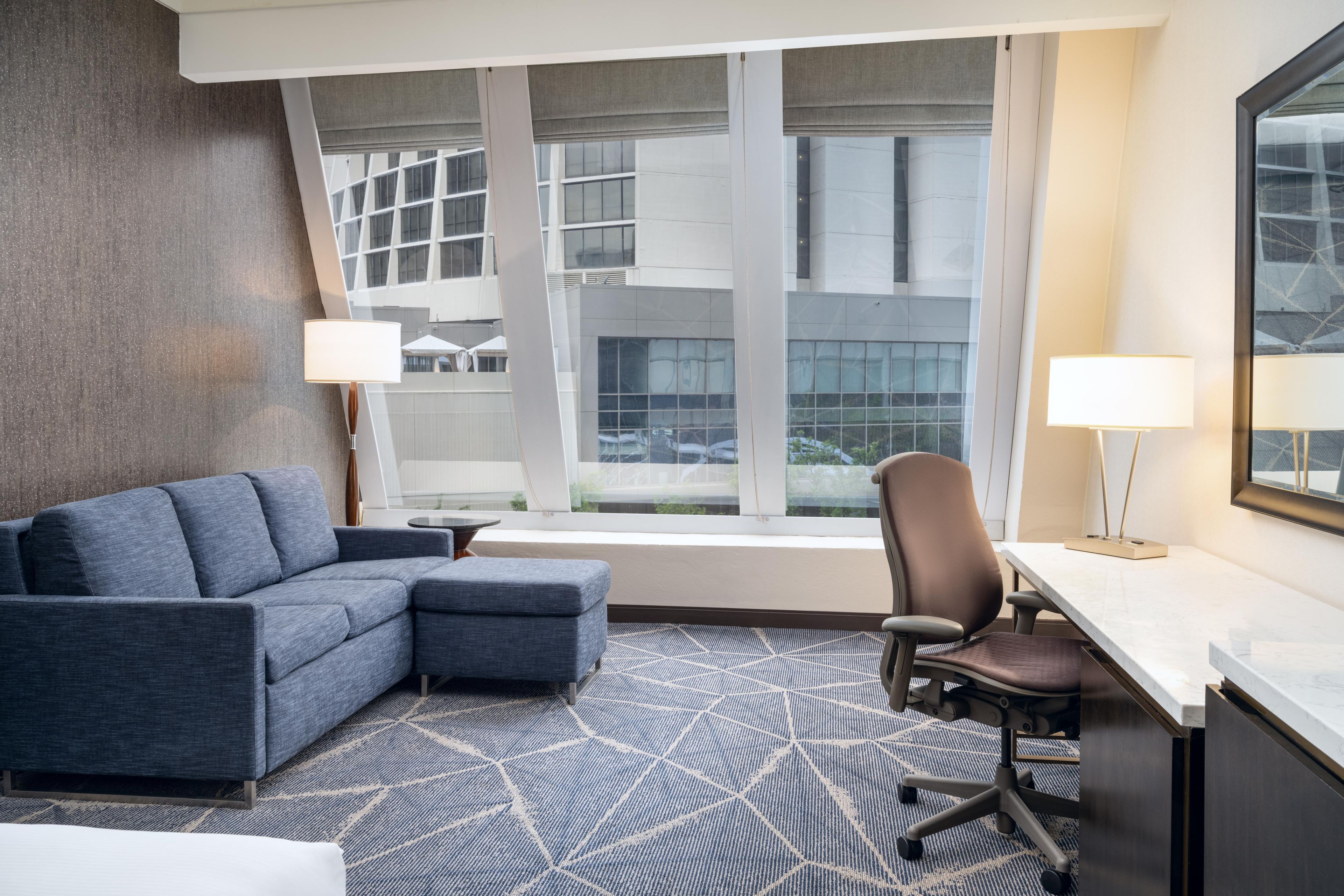 Ein Hotelzimmer im modernen Stil, mit einem grauen modularen Sofa, einem einzelnen Bürostuhl, einem kleinen runden Tisch und einem großen Fenster mit Blick auf ein Gebäude.