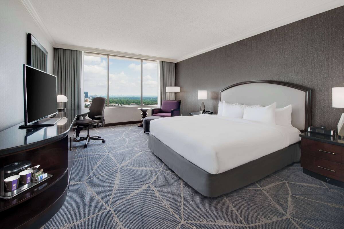 A hotel room with a large bed, a desk with a TV, a chair, and a view of the city.