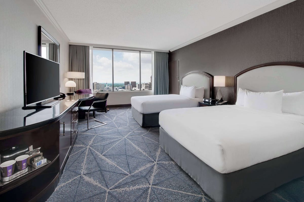A hotel room with a large bed, a smaller bed, a desk with a TV, a chair, and a city view.